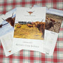 A spiral-bound 2027 wall calendar titled 'Kitchen Coos & Ewes' displayed on a red and green plaid fabric surface. The cover features a highland cow in a grassy field with a tractor, and the pages show monthly calendars with cow photos.